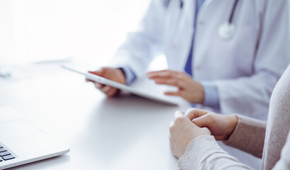 Doctor and patient sitting at the table in clinic while using tablet computer. The focus is on female patient's hands, close up. Medicine concept