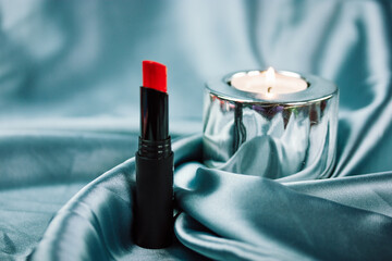 Red lipstick on silver blue silk textile background. Woman's cosmetic table. A burning candle in a...