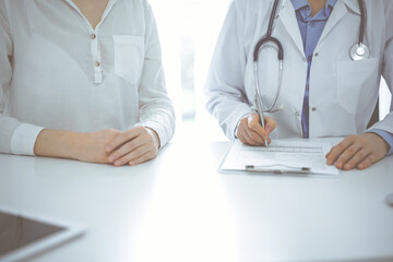 Doctor and patient sitting near of each other at the desk in clinic. The focus is on female physician's hands filling up the medication history record form or checklist, close up. Medicine concept - obrazy, fototapety, plakaty