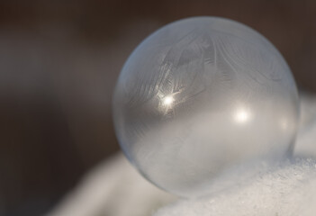 Close-up of a frozen soap bubble lying on fresh snow. The background is brown. The soap bubble is transparent.