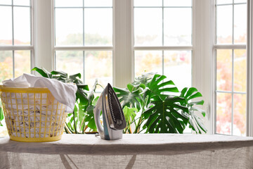 Ironing board with an iron and laundry basket in front of a large window with houseplant
