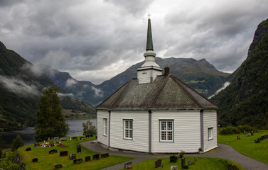 Fototapeta na wymiar The beautiful octagonal Geiranger church next to the cemetery. It is made of white wood with a black slate roof. It has spectacular views of the Geiranger Fjord and the mountains, Norway