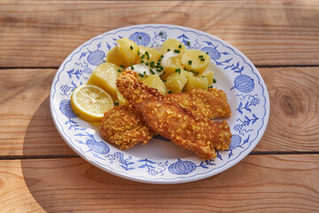 Breaded and deep fried crispy codfish with boiled potatoes, melted butter and chopped fresh chives served on the vintage blue onion porcelain plate on rustic wooden table. Balanced and healthy dish.
