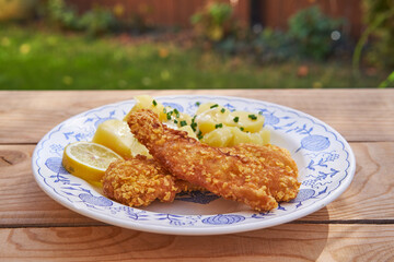 Breaded and deep fried crispy codfish with boiled potatoes, melted butter and chopped fresh chives served on the vintage blue onion porcelain plate on rustic wooden table. Balanced and healthy dish.