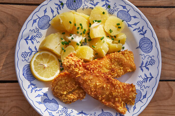 Close up top picture of breaded and deep fried crispy cod fish or haddock fillet with boiled potatoes greased by melted butter and sprinkled by chopped chives. Served on vintage style porcelain plate.