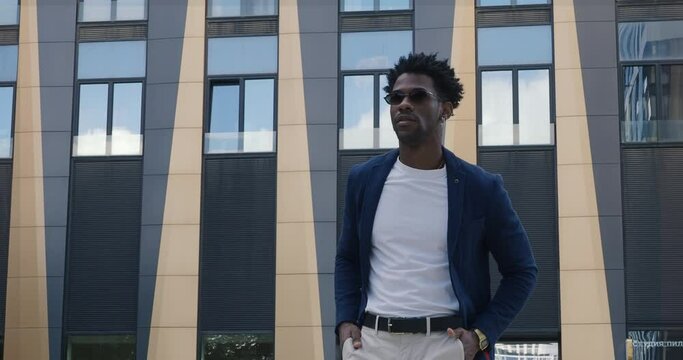 Confident African American male in stylish clothes Blue blazer with T-shirt stands in city next to modern building, looking to side. Portrait Handsome black man in urban locations