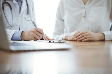 Doctor and patient sitting near each other at the wooden desk in clinic. Female physician's filling up a records form. Medicine concept - obrazy, fototapety, plakaty