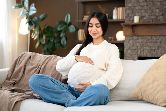 Pregnancy with beautiful pregnant woman hugging her belly sitting on the couch at home. Happy pregnancy with sweet Indian mommy-to-be.