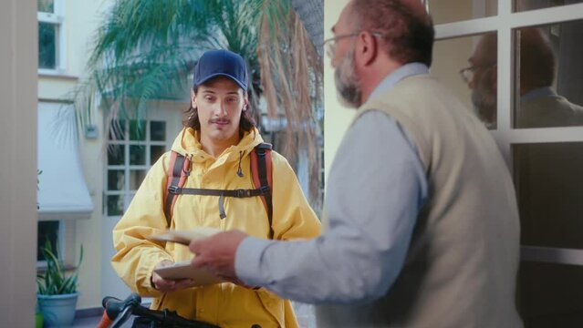 Young male courier in yellow rain jacket giving express mail to senior man standing in the doorway and asking to sign delivery confirmation document