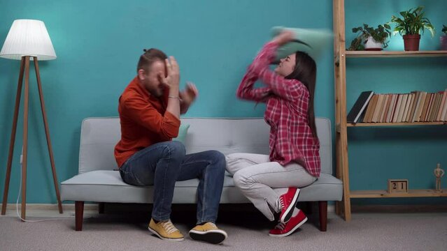 Frustrated young couple are sitting on couch and quarreling with each other, wife hits husband with pillow