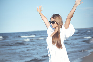 Happy blonde woman is on the ocean beach in a white dress and sunglasses, raising hands