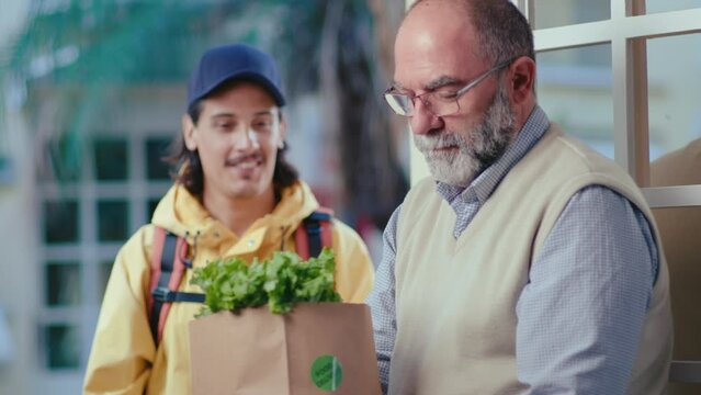 Delivery man with backpack giving paper bag full of groceries to senior man while bringing online order to home. Medium shot, rack focus