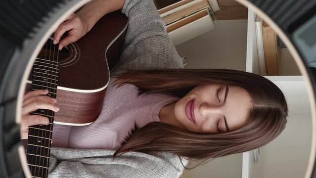 Vertical video. Music vlog. Guitar class. Online tutorial. Cheerful smiling woman playing instrument recording lessons creating content for art blog using ring light home interior.