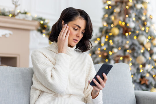 Upset woman sitting alone at home on sofa in living room near Christmas tree, Woman celebrating new year and Christmas, holding phone, received online notification with bad news, unhappy reading