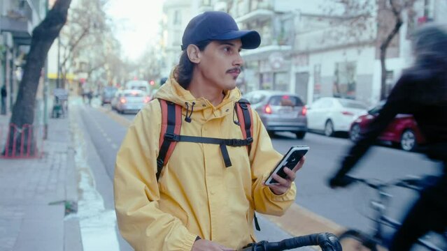Young male courier with backpack standing with bicycle on side of the road, using navigation app on smartphone and looking around