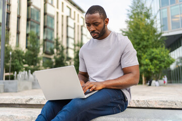 African American man freelancer using laptop typing chatting on urban street in city. Guy having virtual meeting online chat conference. Entrepreneur programmer working on laptop outdoors. Remote work