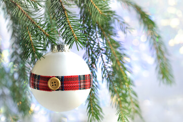 A white Christmas ball with a tartan ribbon hangs on a Christmas fir branch on a shiny white background. Selective focus bokeh with copy space. Christmas card