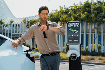 Young man checking time on smartwatch while EV charger to recharging battery from charging station...