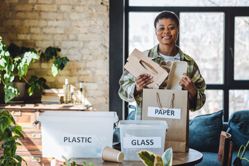 Young African American woman is doing garbage waste sorting at home. Concept of sustainable lifestyle, daily routine to protect planet from pollution, domestic life, simple actions for environment - obrazy, fototapety, plakaty