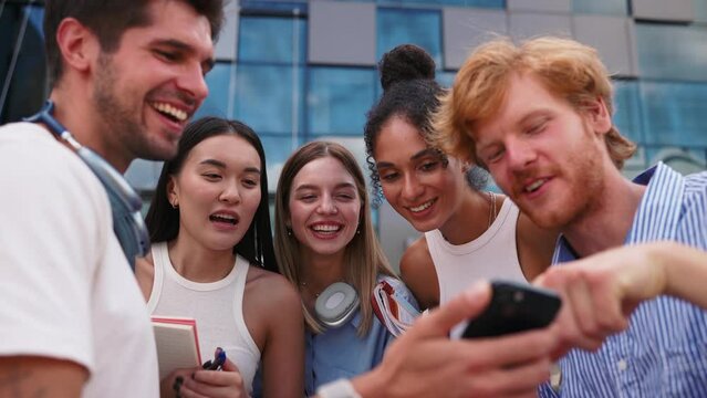 Laughing multinational young students talking and looking at cellphone while standing in circle near university in summertime
