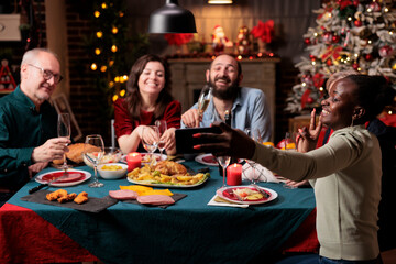People taking pictures at dinner table, making memories of christmas eve festivity gathered around at home. Diverse persons having fun with wine and food, taking photos with smartphone device.