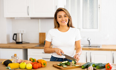 Obraz na płótnie Canvas Cheerful young Hispanic woman following healthy diet preparing vegetable salad for dinner in home kitchen