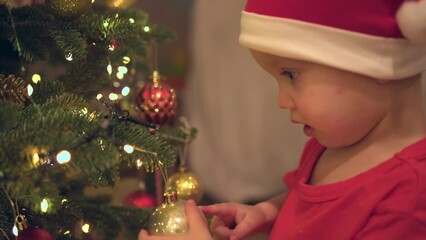 close-up portrait of baby boy in red Santa hat stands near decorated Christmas tree touching golden ball decoration hanging Christmas tree. kid is happy surprised by bright Christmas lights of garland - Powered by Adobe