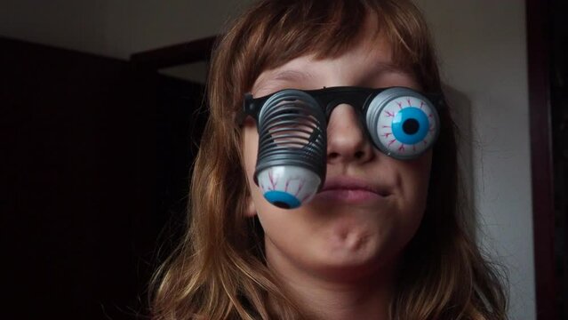 A 9-year-old caucasian girl wearing glasses with eyeballs falling out on springs. A child is preparing for Halloween and trying on funny, scary accessories. Masquerade decoration. Black humor
