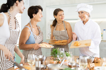 Mature female cook holding cutting board with raw chicken breast in her hands teaching members of cooking course