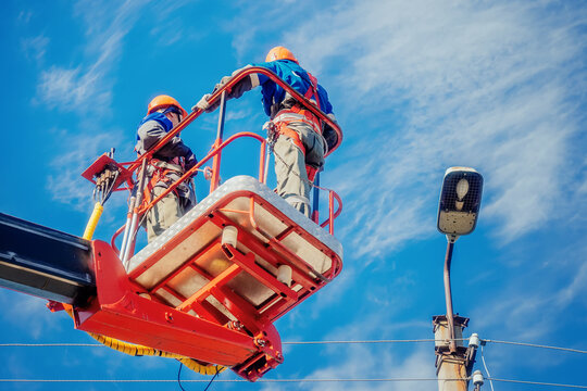 Two electricians from cradle of aerial platform or crane are repairing street lighting lamp. Professional electricians wearing helmets, overalls and insurance work at heights. View of workers from