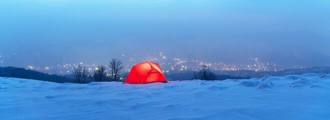 Winter panorama with red tent lighted from the inside against the backdrop of glowing city lights in fog. Tourists camp in winter mountains. Travel concept