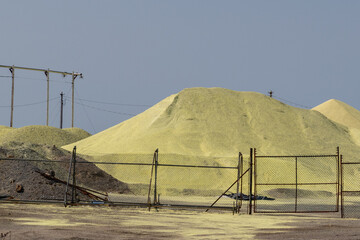 Mounds of yellow sulfur powder behind a metal fence. The pile is used for oil refinement as a waste...
