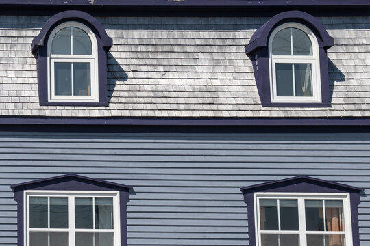 The exterior of a vintage grey painted wooden clapboard house with black trim. The mansard style cedar shingled roof has two arched dormer windows and two casement windows in the historical building. 