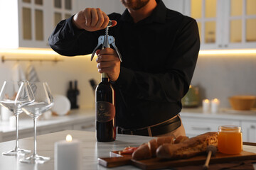 Romantic dinner. Man opening wine bottle with corkscrew at table in kitchen, closeup