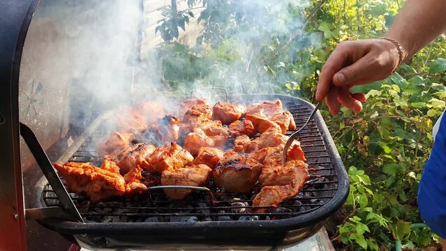 Close up of male hands baking meal outdoors at grill using a fork to turning around meat for not burn out. barbecue and picnic season concept. Feel the great flavour through the dense steam