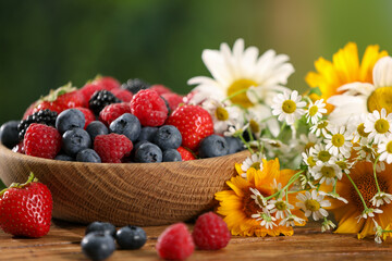 Bowl with different fresh ripe berries and beautiful flowers on wooden table outdoors