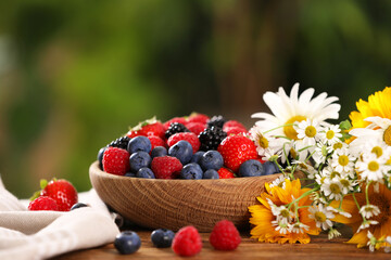 Bowl with different fresh ripe berries and beautiful flowers on table outdoors