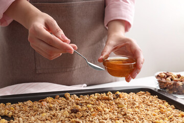 Making granola. Woman adding honey onto baking tray with mixture of oat flakes and other...