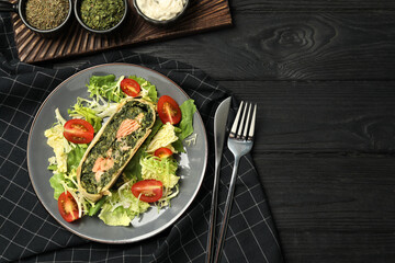 Piece of tasty strudel with salmon, spinach and salad on black wooden table, flat lay. Space for text