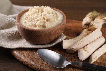 Spicy horseradish sauce in bowl, roots and spoon on wooden table, closeup