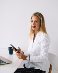Positive watchful female sitting at table messaging on mobile and working on laptop