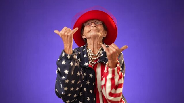 Portrait of shaka hand gesture given by funny crazy grandmother mature woman, 80s, acting wearing US flag jacket isolated on purple background. Concept of cool elderly rock and roll lady.
