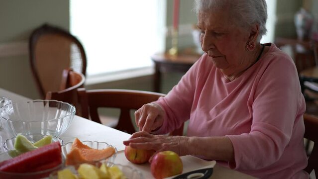 Elderly senior woman sits at table and struggles to cut fruit with a knife.