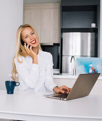 Joyful female sitting at table talking on mobile and typing on laptop