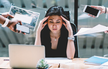 Young disappointed woman feeling headache from deadline surrounded by colleagues at workplace