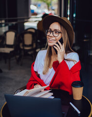 Fototapeta premium Joyful hipster woman in hat talking on smartphone while working at table at cold cafe terrace