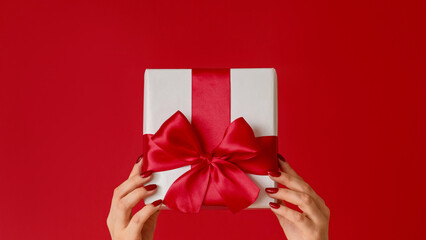 Gift delivery. Christmas surprise. Female hand showing wrapped present in white box with ribbon bow isolated on red copy space background.