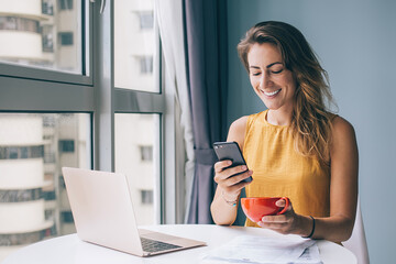 Glad female sitting at table with notebook documents and cup tapping on mobile