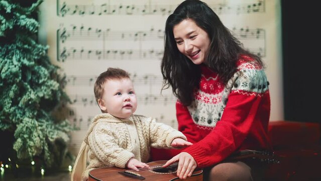 Mom and sweet baby boy in warm red sweaters hug kiss at decorated music sheet notes background and play jingle bells on guitar, celebrate Christmas. Happy family holidays home. Merry Christmas, new ye