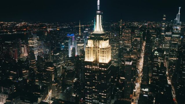 Scenic aerial around the Empire State Building roof highlighted by bright yellow light at night. Outstanding skyscraper on motion background of night New York panorama. Breathtaking Manhattan USA 4K